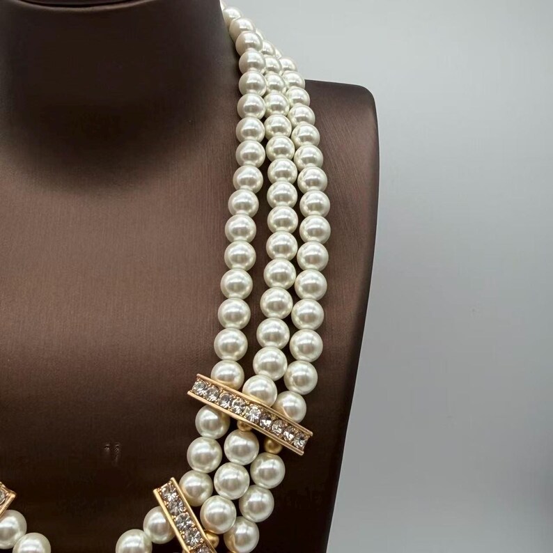 Peut inclure: Un collier multi-rangs compos&eacute; de trois rang&eacute;es de perles blanches uniformes. Les rangs sont reli&eacute;s par deux barrettes rectangulaires en m&eacute;tal dor&eacute;, chacune incrust&eacute;e d'une ligne de petits cristaux clairs et brillants qui ornent le design.