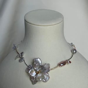 May include: A delicate necklace featuring a large, iridescent flower pendant made of irregularly shaped pearls. The necklace has a gold-coloured chain with smaller pearls and pearl accents in shades of pink and grey.