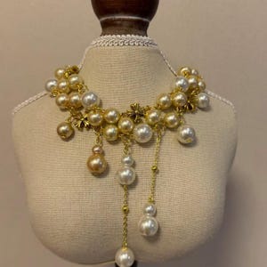 Può includere: Una collana a più fili con perle color crema e ciondoli a croce in metallo color oro. Il design presenta diversi accenti di perle pendenti sospesi da sottili catenelle dorate, creando un aspetto stratificato e asimmetrico con perle di dimensioni varie.