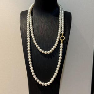 Op de afbeelding: Een lange streng witte parels gerangschikt in een dubbele lus, met een ronde goudkleurige sluiting versierd met kleine doorzichtige steentjes. De ketting bestaat uit uniforme, ronde, glanzende kralen in een continu cirkelvormig ontwerp.