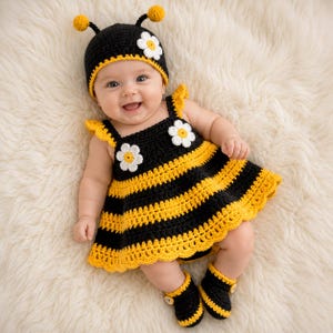 Può includere: Un bambino indossa un costume da ape all'uncinetto fatto a mano, tra cui un vestito a righe nere e gialle, scarpine abbinate e un cappello con antenne e dettagli floreali. Il bambino sorride, sdraiato su una superficie bianca e soffice.
