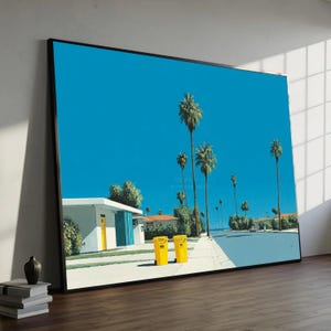 Puede incluir: Una impresión artística enmarcada que representa una escena callejera soleada con palmeras bajo un cielo azul brillante. Dos botes de basura amarillos están en la acera frente a un edificio blanco con una puerta amarilla.