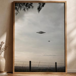 May include: A framed wall art print featuring a gray, disc-shaped unidentified flying object hovering in a cloudy sky above a barbed wire fence and a field, with a small jet aircraft flying nearby and tree branches visible at the top.