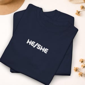 May include: Two navy blue t-shirts are stacked, with the text "HE/SHE" in white block letters. The shirts are on a light background, with a tan hat and dried flowers in the background.
