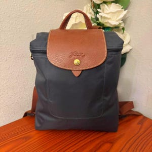 May include: A navy blue backpack with a brown leather flap and handle. The flap features a gold-coloured button and a logo. The backpack has brown straps and a zip closure. The bag is sitting on a wooden surface.