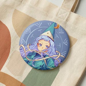 May include: A circular pinback button featuring an anime-style illustration of a character with long, wavy lavender hair wearing a pointed blue hat. The background is a muted blue circle with white line art symbols arranged in a circular pattern.