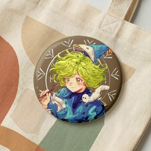 Puede incluir: Un botón redondo con una ilustración de estilo anime de un personaje con cabello verde, vistiendo un atuendo azul y sosteniendo una varita. Un búho blanco está sentado en el hombro del personaje. El botón es marrón con detalles blancos, sobre un bolso tote beige.
