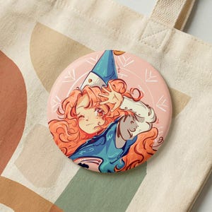 May include: A circular pinback button featuring an anime-style illustration of a character with long, wavy orange hair, wearing a blue pointed hat and a blue garment. The background of the illustration is a soft peach color with white line art accents.