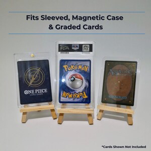 K&ouml;nnte beinhalten: Drei kleine Holzstaffeleien mit Sammelkarten in Schutzh&uuml;llen. Der Text oben lautet Fits Sleeved, Magnetic Case & Graded Cards. Der Text unten lautet *Cards Shown Not Included. Die H&uuml;llen enthalten One Piece, Pokemon und Magic Karten.