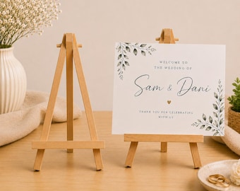 Könnte beinhalten: Zwei helle Holzstaffeleien, von denen eine ein weißes, rechteckiges Hochzeitsschild mit grünen Blattillustrationen und dem Text: Welcome to the wedding of Sam & Dani, thank you for celebrating with us, sowie einem kleinen goldenen Herz trägt.