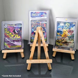 May include: Three miniature wooden easels, each holding a collectible card in a clear protective case. The cards feature vibrant illustrations and Japanese text. The central card is graded and labelled "MEGA CHARIZARD EX". The text "*Cards Not Included" is at the bottom.