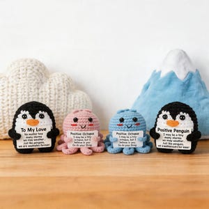 May include: Four crocheted stuffed animals: two penguins and two octopuses. The penguins are black and white, and the octopuses are pink and blue. Each animal holds a small white sign with text. A cloud and mountain plush are in the background.