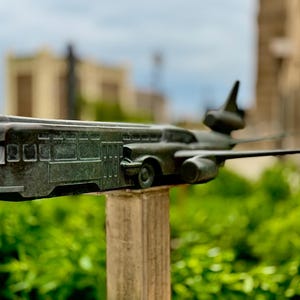 Può includere: Una scultura color bronzo di un tram con ali e timoni di coda di aereo. La scultura è montata su un palo di legno, con uno sfondo verde sfocato di fogliame ed edifici.