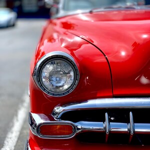 Może przedstawiać: Zbliżenie klasycznego, czerwonego zabytkowego samochodu, prezentujące jego błyszczący lakier i chromowane detale. Samochód ma okrągły reflektor z chromowaną ramką i chromowany zderzak. Maska i przód samochodu są ostre.