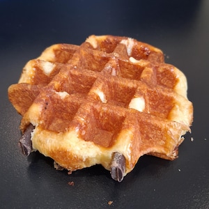 May include: A golden-brown waffle with a crispy exterior and a soft interior. The waffle has a grid pattern and is studded with dark chocolate pieces. The image is a close-up shot, highlighting the texture and details of the baked treat.