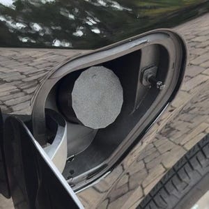 May include: A dark-colored vehicle fuel filler door is open, revealing a circular, textured, light gray fuel cap. The surrounding body panel is glossy and reflective, showing a distorted view of a paved surface. A small metal latch mechanism is visible.