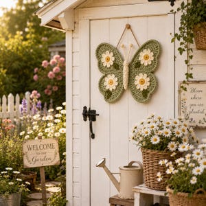 May include: A crochet butterfly wall hanging featuring sage green wings decorated with four white daisy motifs with yellow centers. The butterfly has a cream-colored body and antennae, and it is suspended by a thin brown string against a white door.