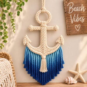 Puede incluir: Un tapiz de macramé con forma de ancla, que presenta una cuerda anudada de color blanco roto en la parte superior y flecos con degradado azul en la parte inferior. El diseño del ancla incluye un anillo superior circular y dos brazos curvos.