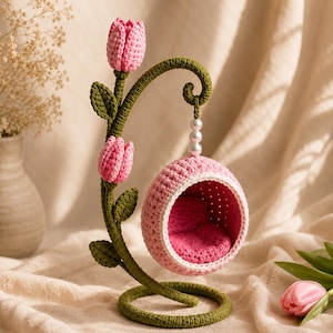 May include: A crochet hanging chair suspended from a green stem featuring two pink tulip blossoms and green leaves. The chair is round, light pink with a dark pink interior, and hangs from a curved stem by a string of three white beads.