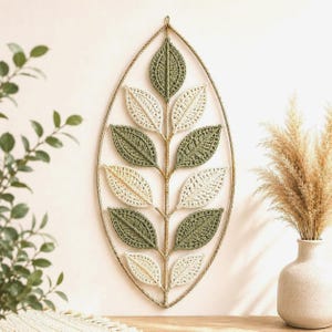 May include: A wall hanging featuring a pointed oval frame made of twisted cord, containing a central stem with alternating crocheted leaves in shades of sage green and cream. The leaves are arranged symmetrically along the vertical axis.