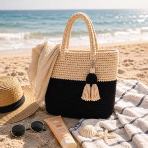 Op de afbeelding: Een tweekleurige gehaakte tote bag met een crèmekleurig bovendeel en een zwart onderdeel, geaccentueerd met een crèmekleurige kwastjesbedel aan een handvat. De tas wordt vergezeld door een lichtbeige sjaal die over de zijkant gedrapeerd is.