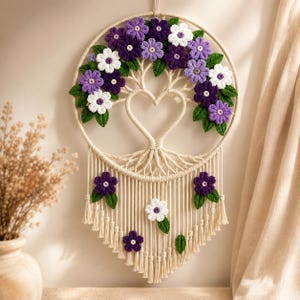 May include: A macrame wall hanging featuring a circular frame with a central heart-shaped tree design. The tree is adorned with crocheted flowers in shades of purple, lavender, and white, accented by green leaves. Long fringe hangs below.