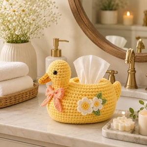 May include: A yellow crochet tissue box cover shaped like a duck, featuring a peach-coloured bow around its neck and two white daisy appliques with green leaves on its side. The duck has a small orange beak and black bead eyes for detail.