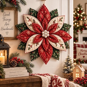 May include: A quilted fabric poinsettia wall hanging featuring layered red, cream, and dark green petals with gold snowflake and holly berry patterns. The centre is adorned with a cluster of six white pearls, and the piece hangs from a twine loop.
