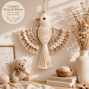 May include: A cream and tan crochet bird wall hanging with layered scallop wings and a long tassel tail. The bird features a small orange beak and a black bead eye. The design includes a hanging loop at the top for display on a wall surface.