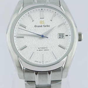 May include: A silver-coloured Grand Seiko automatic wristwatch with a textured dial and a date display at the 3 o'clock position. The watch features a silver bracelet and the text "Grand Seiko" and "AUTOMATIC HI-BEAT 36000" on the dial.