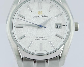 May include: A silver-toned Grand Seiko automatic wristwatch with a textured dial and a date display at the 3 o'clock position. The watch features a silver bracelet and the text "Grand Seiko" and "AUTOMATIC HI-BEAT 36000" on the dial.