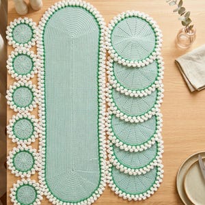 Pode incluir: Um caminho de mesa e bases para copos de crochê a condizer, em verde suave. O caminho de mesa é oval e as bases para copos são redondas. Ambos têm uma borda branca texturizada. O conjunto é exibido numa mesa de madeira.
