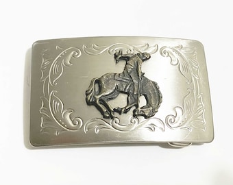 May include: Silver-toned rectangular belt buckle with a bronze-colored cowboy riding a bucking bronco. The buckle has an ornate floral design around the edges.