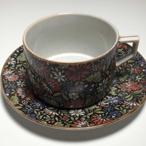 May include: A floral teacup and saucer set. The cup and saucer feature a black background with a pattern of colorful flowers in shades of red, white, and blue. The rims are trimmed with gold. The teacup has a handle.