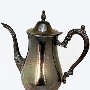 Puede incluir: Una cafetera plateada con detalles ornamentados. Presenta un pico curvo, un asa decorativa y una tapa con un pequeño adorno. La cafetera se apoya sobre una base decorativa con tres patas. El diseño es vintage y elegante.