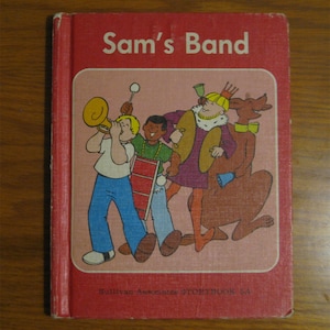 Könnte beinhalten: Ein rotes Hardcover-Buch mit dem Titel Sam's Band und einer zentralen Illustration von Zeichentrickfiguren, die Musikinstrumente wie eine Trompete, eine Trommel und Becken spielen, mit dem Text Sullivan Associates STORYBOOK 5A am unteren Rand.