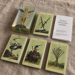 May include: A Pagan Otherworlds Tarot deck set featuring a holographic box, a general instruction booklet, and five tarot cards including Death, The Emperor, and Two of Swords, all displayed with pale green backgrounds and detailed illustrations.