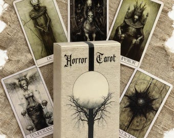 May include: A deck of "Horror Tarot" cards with a beige box featuring a black tree and a full moon design. The cards depict gothic illustrations of skeletons, figures, and symbols. The cards are arranged around the box on a textured, beige surface.
