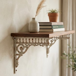 May include: A wooden shelf with ornate, white metal brackets is mounted on a wall. The shelf holds a small ceramic vase with a dried plant, a stack of books, and a potted plant. The shelf has a dark wood finish.