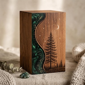 May include: A rectangular wooden urn featuring a vertical, wavy inlay of dark green resin with gold flecks. The wood surface displays a laser-engraved forest scene with a tall pine tree, smaller trees in the distance, a crescent moon, and stars.