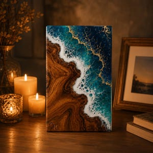May include: A rectangular wooden art piece with a resin ocean design. The artwork features a brown wooden base, white foamy waves, and a teal and navy blue ocean with gold accents. The piece is displayed on a wooden surface with candles and a framed picture.