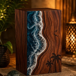 May include: A rectangular wooden urn featuring a dark brown wood grain finish. One side displays a decorative resin inlay depicting a blue ocean wave with white sea foam. The wooden surface includes engraved silhouettes of two palm trees and birds.
