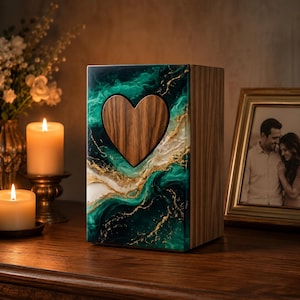 May include: A wooden urn with a heart-shaped cutout. The front panel features a swirling design of green, gold, and white resin. The sides are made of wood. The urn is displayed on a wooden surface with lit candles and a framed photo in the background.