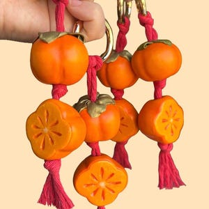 May include: A collection of orange persimmon ornaments with gold-colored accents and red string. Some persimmons are whole, while others are cut to reveal a star-shaped design. The ornaments are attached to gold-colored rings.