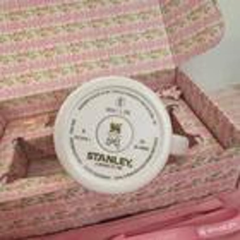 Puede incluir: Un flex&oacute;metro Stanley blanco con texto negro y un logotipo, dentro de una caja rosa floral. El flex&oacute;metro tiene forma redonda con un asa. La caja est&aacute; abierta, revelando el flex&oacute;metro y su embalaje.