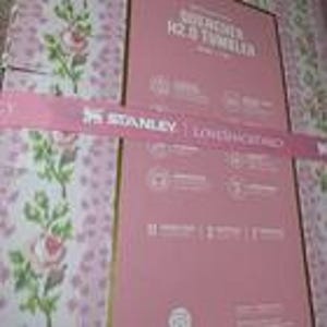 Puede incluir: Caja rosa para un vaso Stanley Quencher H2.0 con una cinta rosa. La caja presenta el logotipo de Stanley y los detalles del producto. El fondo muestra un estampado floral con rosas rosas y verdes.