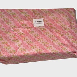 Puede incluir: Un paquete rectangular envuelto en papel con estampado floral rosa. El papel presenta un dise&ntilde;o repetido de rosas rosas y hojas verdes sobre un fondo de rayas diagonales. Una etiqueta blanca con el texto "STANLEY LOVESHACKFANCY" est&aacute; adherida.