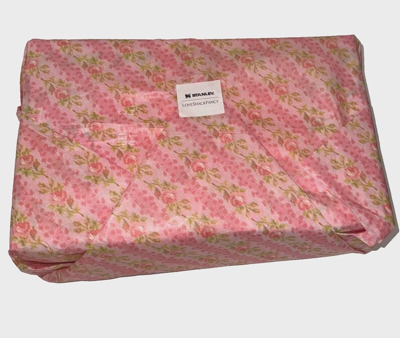 Puede incluir: Un paquete rectangular envuelto en papel con estampado floral rosa. El papel presenta un dise&ntilde;o repetido de rosas rosas y hojas verdes sobre un fondo de rayas diagonales. Una etiqueta blanca con el texto "STANLEY LOVESHACKFANCY" est&aacute; adherida.
