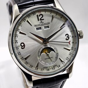May include: A silver-toned analogue wristwatch with a black leather strap. The dial features a sunburst finish, Arabic numerals at 3, 9, and 12, a moon phase indicator, a date calendar ring, and the text Jaeger-LeCoultre, 2027, JAN, and SWISS MADE.