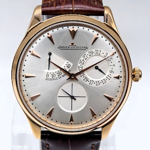 May include: A gold-tone wristwatch with a silver dial, featuring the Jaeger-LeCoultre logo, a power reserve indicator, a date sub-dial, and a small seconds sub-dial. The watch has rose gold-tone hour markers and a brown leather strap with a textured finish.